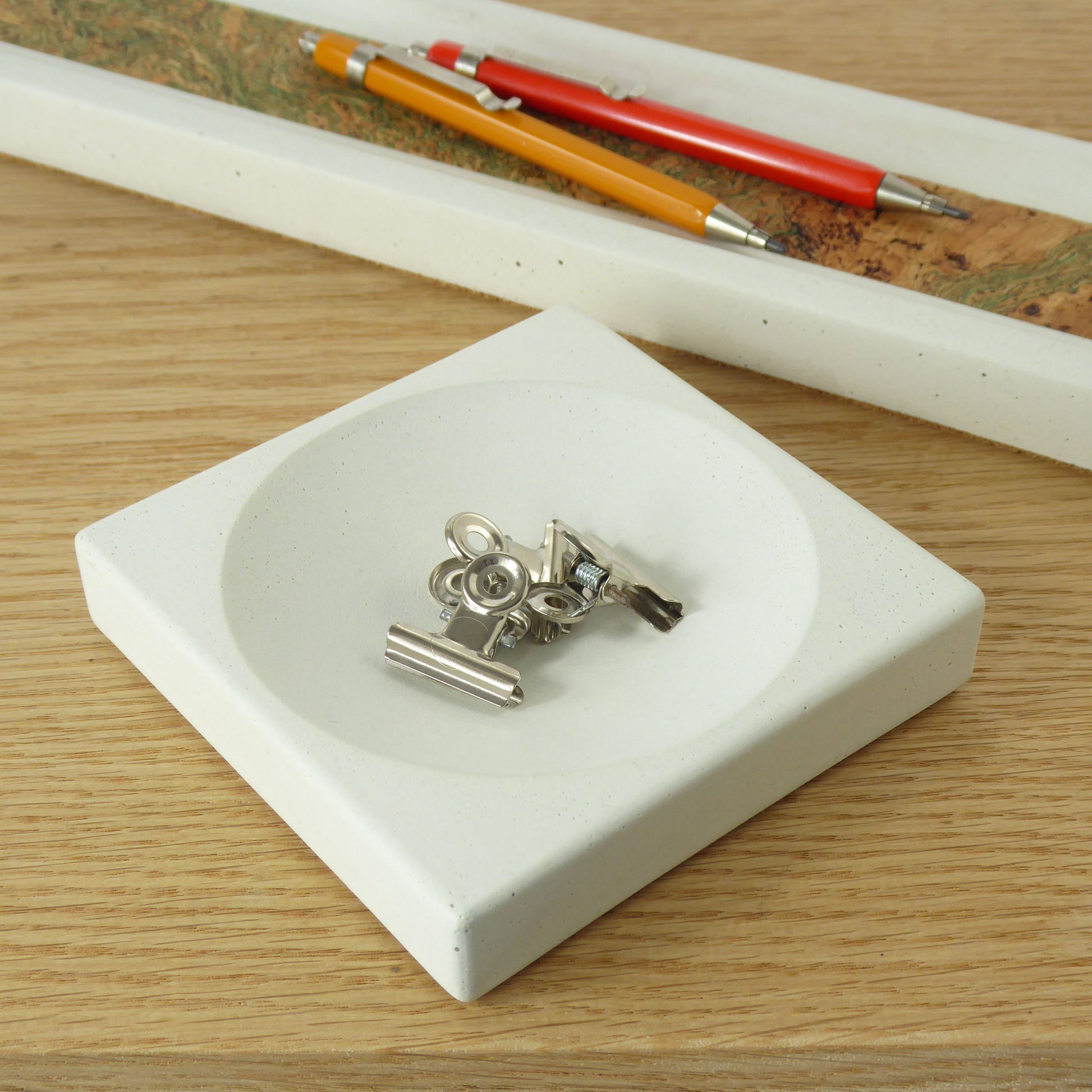 Metal stationery clips in a white concrete tray with a pen tray in the background on a wooden desk