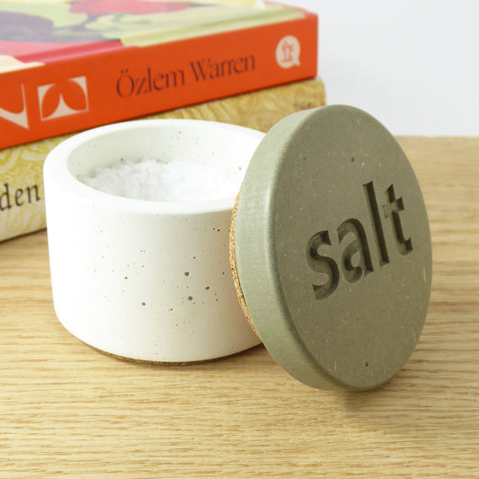 White concrete salt container with a sage coloured wooden lid engraved with the word 'salt' on a wooden kitchen worktop next to cookbooks