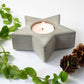 Concrete star-shaped candle holder with a lit candle, surrounded by greenery and pine cones on a white background.
